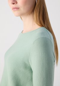 Világoszöld kötött pulóver, lekerekített nyakrésszel és bordázott textúrával, amely kiemeli a puha anyagát és a testhez simuló fazonját.