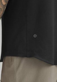 T-shirt de sport noir avec une surface texturée ; présente un logo subtil sur l'ourlet. Associé à un pantalon beige clair, mettant en valeur une coupe décontractée.
