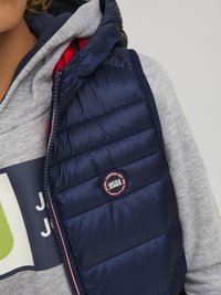 Námornícko modrá prešívaná vesta s červeným podšívaním, s horizontálne prešívanými panelmi a logom. Nosená cez šedú mikinu.
