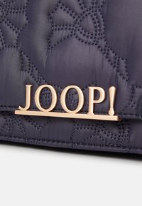 Borsa in tessuto quilted blu navy con placca logo "JOOP!" in tonalità oro, che mostra intricati motivi di cucitura sulla superficie.