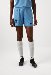 Ljusblå fotbollsshorts i andningsbart material, med en svart lejonemblem och Nike-logotyp, matchade med vita knästrumpor och svarta fotbollsskor.