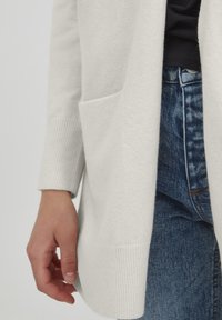 Langer, hellgrauer Cardigan aus weichem Stoff, mit gerippten Bündchen und einer Fronttasche, getragen über Jeans.