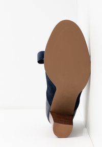 Vue de la semelle d'une botte cheville en daim bleu marine avec une semelle en caoutchouc marron et un talon en bois massif sur fond blanc.