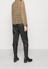 Blumenmuster-Bluse mit langen Ärmeln in Schwarz, Gelb und Orange; kombiniert mit schwarzen Leder-Hosen in schmaler Passform und kniehohen schwarzen Stiefeln mit breiten Absätzen.