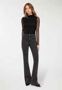 Donna in piedi che indossa una maglia nera trasparente a maniche lunghe, jeans a zampa d'elefante grigio scuro a vita alta e scarpe nere con punte appuntite e fiocchi borchiati.