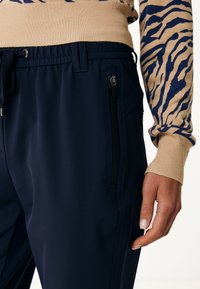 Pantalones azul marino con un acabado texturizado, que presentan una cintura elástica y un bolsillo lateral con cremallera. La mano del modelo descansa cerca del bolsillo.