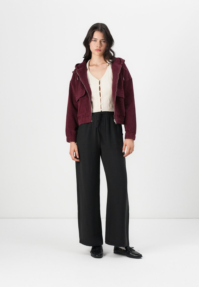 Burgundowa kurtka z kapturem, krótka, beżowa bluzka z guzikami oraz czarne spodnie szerokie. Gładkie faktury z luźnym krojem.