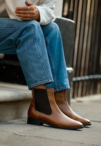 Bottes cheville en cuir marron avec un bout pointu, des panneaux latéraux élastiques et un petit talon noir, associées à un jean bleu clair et un pull blanc.