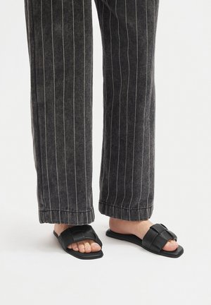 Czarne klapki na płaskiej podeszwie z plecionym paskiem, zestawione z ciemnymi, szerokimi spodniami w prążki, prezentujące gładką, matową fakturę.