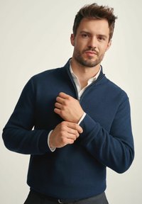 Granatowy sweter z półka z suwakem, wykonany z dzianiny o teksturowanej fakturze oraz z prążkowanymi mankietami. Nosi się go na białej koszuli z kołnierzykiem, z zamkiem błyskawicznym i nowoczesnym krojem.