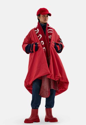 Personne portant un manteau rouge surdimensionné, une écharpe rouge, une casquette rouge, des bottes rouges, un pull bleu marine avec des rayures rouges, et un pantalon bleu foncé sur un fond uni.