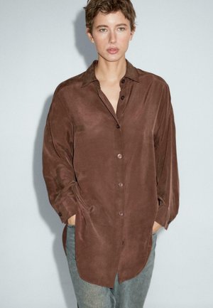 Camicia marrone a maniche lunghe con una trama liscia, chiusura frontale a bottoni e tasche laterali. Indossata sopra jeans azzurri chiari.