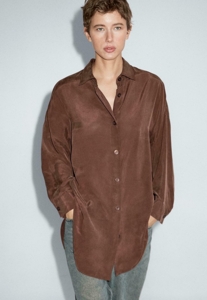 Camicia marrone a maniche lunghe con una trama liscia, chiusura frontale a bottoni e tasche laterali. Indossata sopra jeans azzurri chiari.