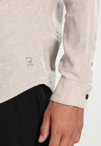 Beżowa lniana koszula z długimi rękawami, zaokrąglonym dołem i brązowymi guzikami na mankietach, z małą markową metką po boku.