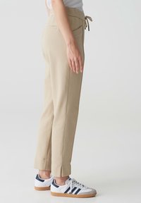 Personne portant un pantalon beige à cordon et des baskets blanches avec des rayures bleu marine, debout de profil sur un fond clair uni.