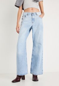 Ανοιχτό μπλε τζιν παντελόνι με φαρδιά μπατζάκια, κατασκευασμένο από ντένιμ, με ψηλή μέση, τέσσερις τσέπες και κλείσιμο με κουμπί, συνδυασμένο με σκούρα μπορντό μπότες με χοντρό τακούνι.