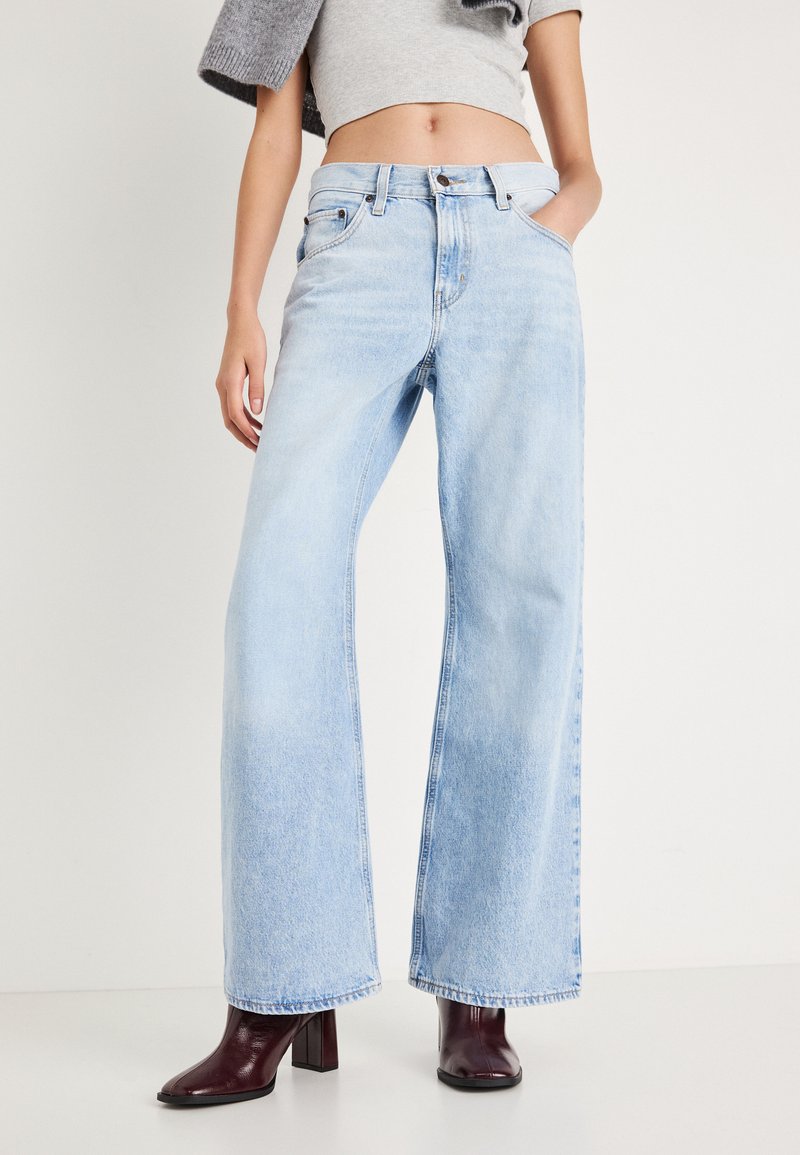 Ανοιχτό μπλε τζιν παντελόνι με φαρδιά μπατζάκια, κατασκευασμένο από ντένιμ, με ψηλή μέση, τέσσερις τσέπες και κλείσιμο με κουμπί, συνδυασμένο με σκούρα μπορντό μπότες με χοντρό τακούνι.