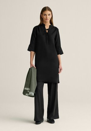 Kvinde iført sort lang tunika med bindebånd ved halsen, sorte bukser med vide ben, sorte sko, og holder et foldet grønt tørklæde i den ene hånd.