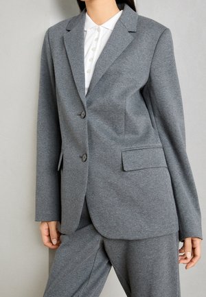 Vrouw draagt een grijs colbert met twee knopen en bijpassende broek over een wit poloshirt met knopen, staand tegen een effen lichte achtergrond.