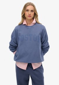 Sudadera azul con la impresión "BOSTON", cuello redondo y hombros caídos, superpuesta a una camisa de collar rosa, combinada con pantalones oscuros.
