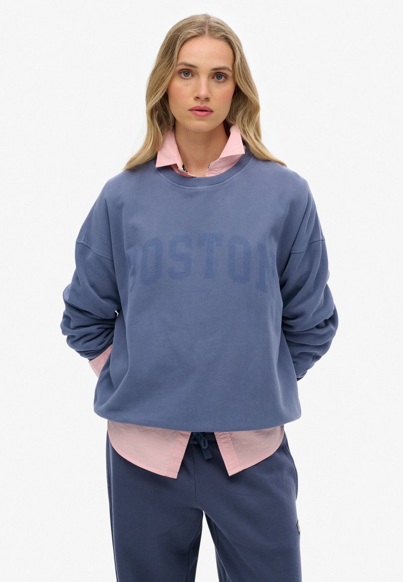Sudadera azul con la impresión "BOSTON", cuello redondo y hombros caídos, superpuesta a una camisa de collar rosa, combinada con pantalones oscuros.