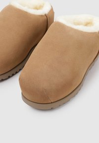 Pantofole in suede marrone con una morbida fodera in pelliccia bianca, design a punta arrotondata e suola in gomma testurizzata per una migliore presa. Finitura esterna liscia.