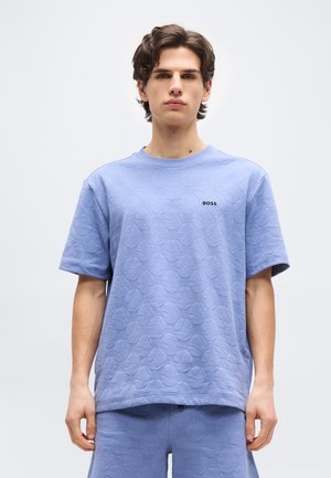 Mlad moški stoji, oblečen v svetlo modro teksturirano srajco s kratkimi rokavi in ujemajoče kratke hlače z logotipom "BOSS" na prsih.