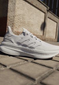 Scarpa da corsa Adidas Ultraboost bianca con tomaia in maglia strutturata e suola spessa ammortizzata su pavimento esterno con sfondo di muro in mattoni.