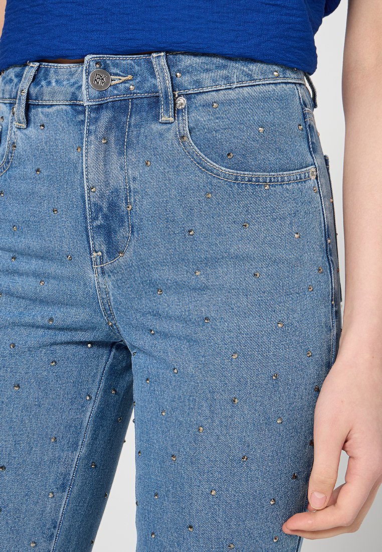 Vaqueros de mezclilla azul de cintura alta, con remaches metálicos dispersos como adorno. Diseño clásico de cinco bolsillos con cierre de botón.