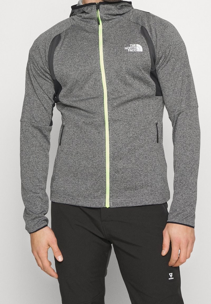 Giacca grigia con zip e cappuccio, con cerniera verde neon, inserti laterali neri e due tasche laterali, realizzata in materiale leggero e strutturato.