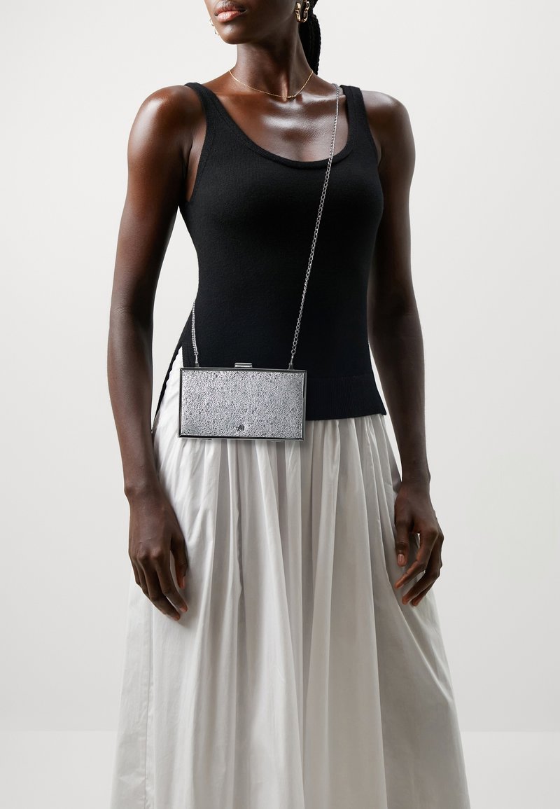 Stříbrná obdélníková kabelka s texturovaným povrchem, řetízkem a zavíráním na klips, v kombinaci s černým topem bez rukávů a bílou plisovanou sukní.