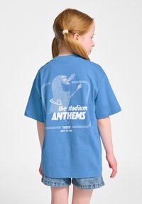 Lichtblauwe t-shirt met korte mouwen, met een afbeelding van een karakter op de achterkant en de tekst "de stadion ANTHEMS" in vette witte letters.