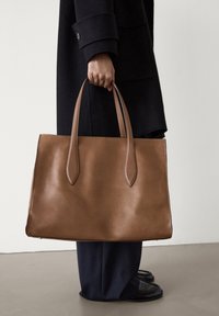 Sac porté épaule en cuir marron avec des poignées allongées, texture lisse et design minimaliste. Tenue par une personne en tenue sombre.
