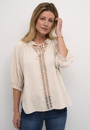 Light beige blouse with a round neckline, tie closure, puffed sleeves, and intricate lace detailing down the front. Soft fabric texture.
