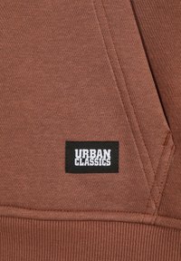 Tejido de sudadera marrón con textura visible, que presenta una etiqueta de logo negra de Urban Classics. Detalle de costura en los bordes. Diseño suave y casual.