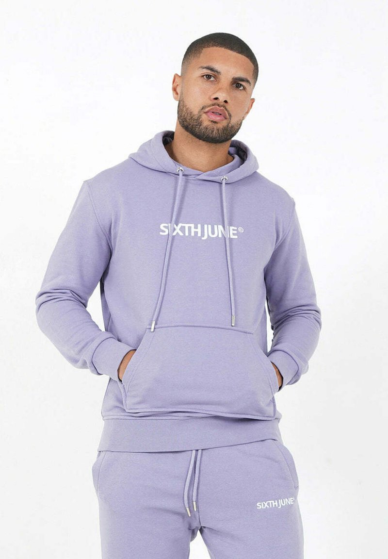 Hombre vestido con un conjunto de sudadera y pantalones de chándal en color lavanda con el logo "SIXTH JUNE", con las manos en el bolsillo de la sudadera, frente a un fondo blanco liso.