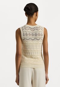 Kvinna med håret i en bulle iförd ärmlös beige virkad topp och krämfärgade byxor, sedd bakifrån mot en enkel vit bakgrund.