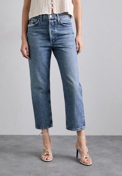 Jeans in denim blu chiaro a gamba dritta con vita alta, caratterizzati da un bottone frontale e leggeri segni di usura. Abbinati a sandali con tacco alto e cinturini.
