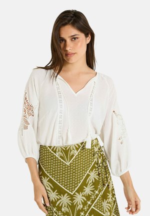 Blusa blanca con mangas abullonadas, detalles de encaje y escote en V. Combinada con una falda verde con estampado de palmeras.