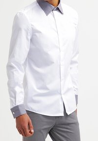 Camicia bianca a maniche lunghe con colletto e polsini grigi, caratterizzata da una texture liscia e chiusura con bottoni davanti.