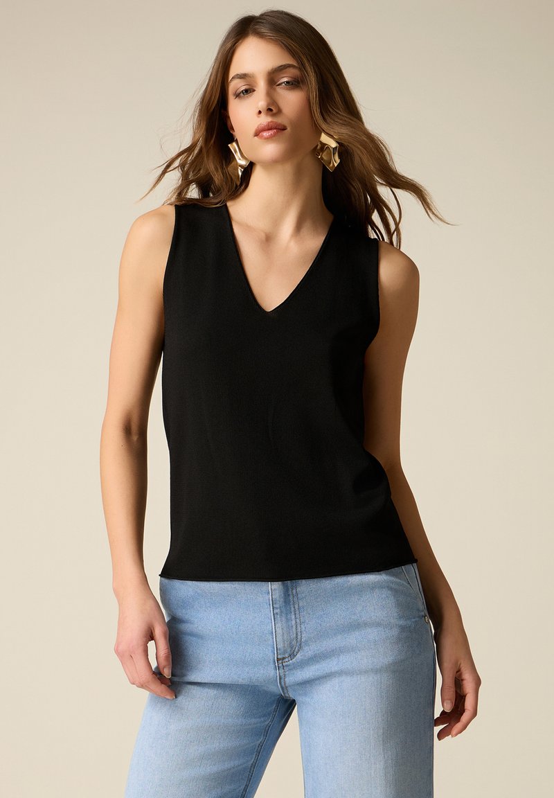 Donna con top nero senza maniche a scollo a V, jeans azzurri chiari e grandi orecchini geometrici dorati, in piedi davanti a uno sfondo beige.