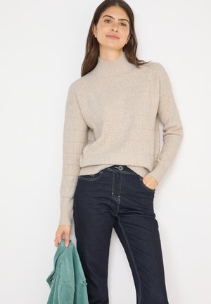 Beige gestrickter Pullover mit hohem Kragen, der eine gerippte Textur und helle Streifen aufweist, kombiniert mit dunkelblauen Jeans und einer türkisfarbenen Jacke in der Hand.