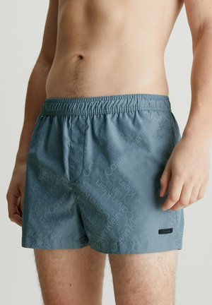 Pantaloni da mare blu con cintura elastica, con un motivo a logo ripetuto e una piccola etichetta nera sul lato sinistro inferiore.