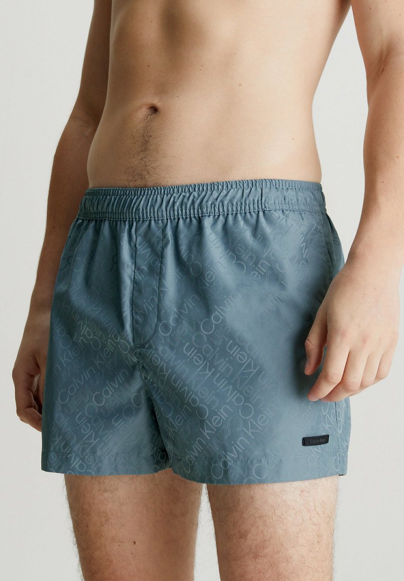 Pantaloni da mare blu con cintura elastica, con un motivo a logo ripetuto e una piccola etichetta nera sul lato sinistro inferiore.