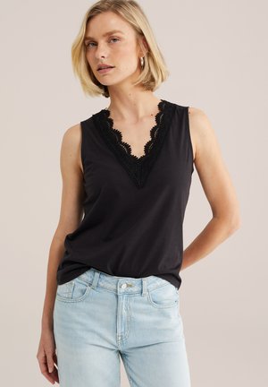 Femme portant un haut noir sans manches avec un décolleté en V en dentelle et un jean bleu clair, posant avec une main derrière le dos devant un fond uni.