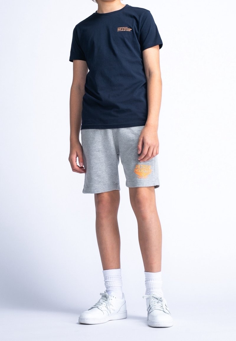 Enfant portant un t-shirt bleu marine, un short gris avec un logo orange sur la jambe gauche, des chaussettes blanches et des baskets blanches, debout sur fond blanc.