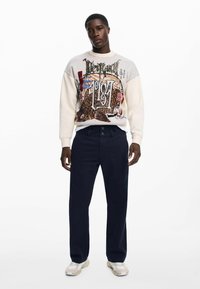 Maglione con stampa grafica in crema e marrone, abbinato a pantaloni scuri e sneaker crema. Il motivo distintivo include testo e disegni.