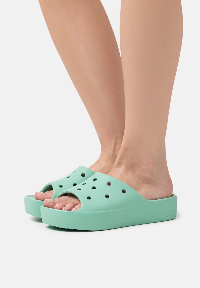 Pieds portant des sandales compensées vert clair à enfiler avec des trous de ventilation ronds sur un fond clair uni.