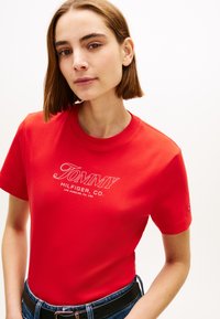 Κόκκινο βαμβακερό μπλουζάκι με λευκό λογότυπο "TOMMY HILFIGER CO." με καλλιγραφική γραφή και κλασικό στρογγυλό ντεκολτέ. Συνδυάζεται με μαύρη ζώνη και τζιν παντελόνι.
