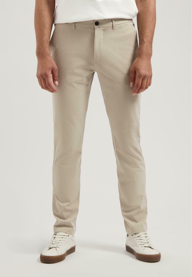 Muž oblečený v béžových slim-fit nohaviciach a bielych teniskách stojí na čistom svetlom pozadí.